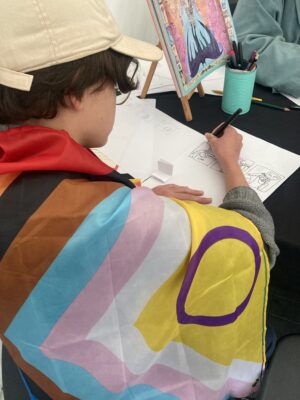 En person med mörkt hår och ljus keps med en pride-flagga draperad över axlarna fotad snett bakifrån och ovanifrån. I bilen syns bordet där personen sitter och tecknar en serie